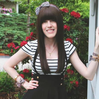 A young white woman with long brown hair and bangs over her eyes. She is wearing a short-sleeved white-and-black striped shirt and black pants with suspenders. Her left arm is leaning against a wall. Behind her is greenery with red flowers.