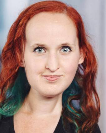 Headshot photo of a white woman with red and teal hair, wearing a black v-neck t-shirt.