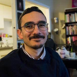 A head and shoulders shot of Angelo, facing the camera and half-smiling. He has short dark hair, a short moustache and goatee, and round metal eyeglasses, and is wearing a navy sweater and striped collared shirt. He is sitting in front of a bookshelf, kitchen, and standing lamp.