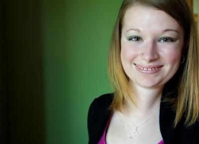 Photo of a room with a green colored wall in the background. On the right-side of the photo is a young white woman with shoulder-length blonde hair. She is wearing a black cardigan and a scoop neck purple top. She is smiling at the camera.