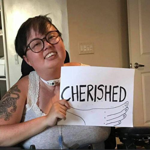 A photo of Stacey, a mixed race Korean and white queer person, holding a paper sign that reads “Cherished.” The photo is stylistically torn at the edges with cursive text appearing across the bottom, “Stacey Milbern.”