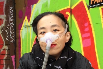 Asian American woman in a wheelchair. She is wearing a black jacket with a black patterned scarf. She is wearing a mask over her nose with a tube for her Bi-Pap machine. Behind her is a wall full of colorful graffiti
