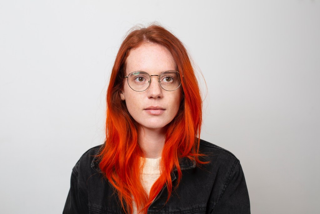 Color photograph of Beatrice Adler-Bolton, simple portrait photo of a pale, freckled white woman with natural red to dyed orange long wavy hair, wearing round gold rimmed glasses and a black t-shirt in front of a white background.