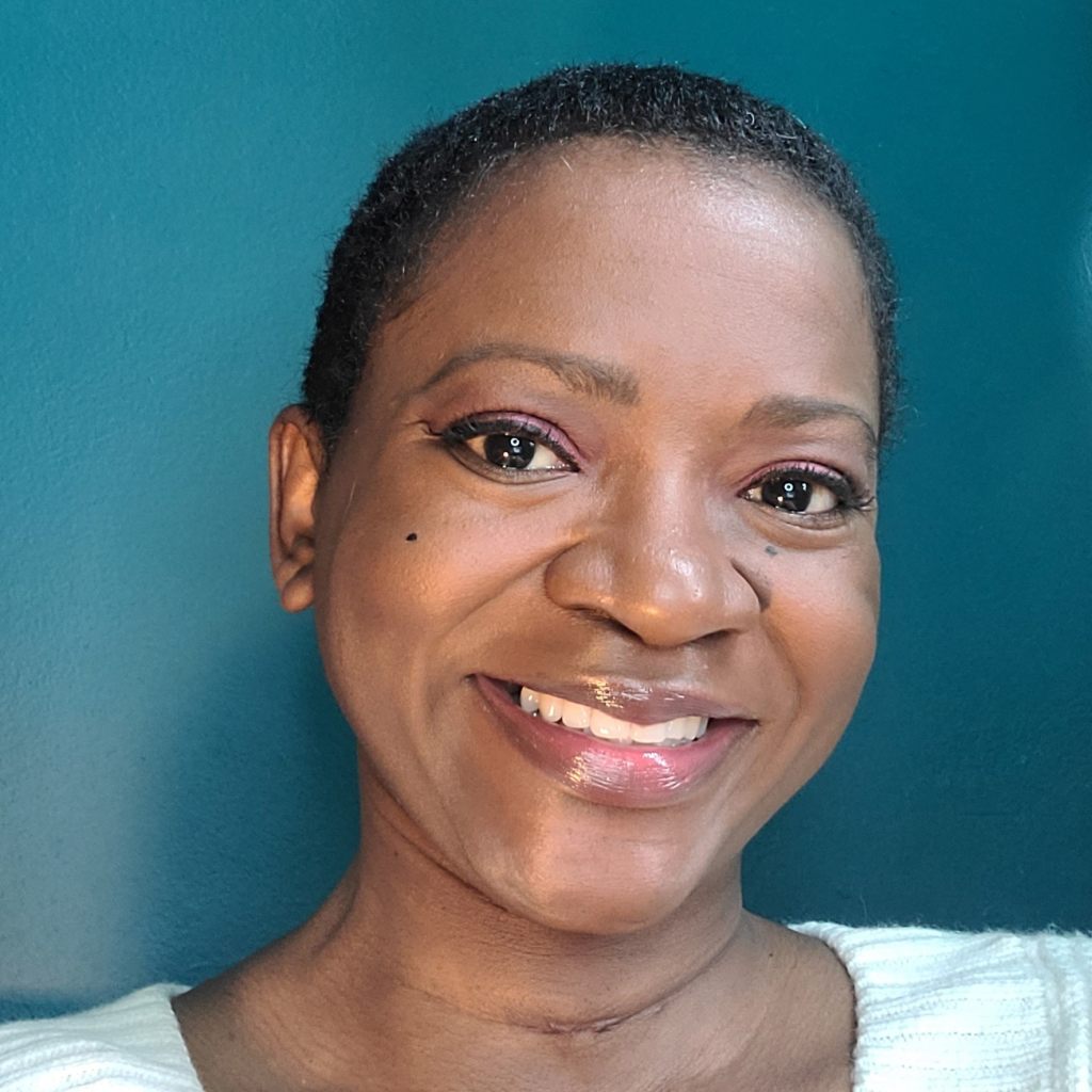 Photo of Carolyn Hinds, a Black female with dark brown complexion, close cropped black hair, wearing a wide smile, and white sweater, in front of a teal colored wall.