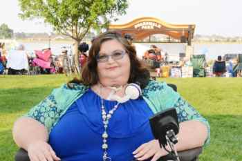 Carrie Ann Lucas, a white woman with long brown hair wearing glasses, a blue top along with a green patterned cardigan and necklace. She has a tube attached to her neck connected to a ventilator. She is sitting in a power chair with in front of a grassy park .