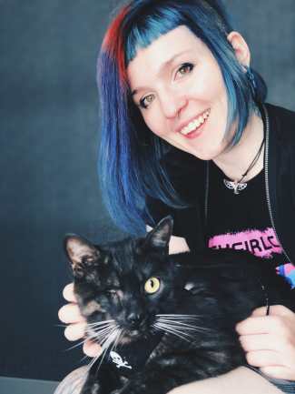 Cherry Smiling at the camera with their one eyed cat Odin on their lap.