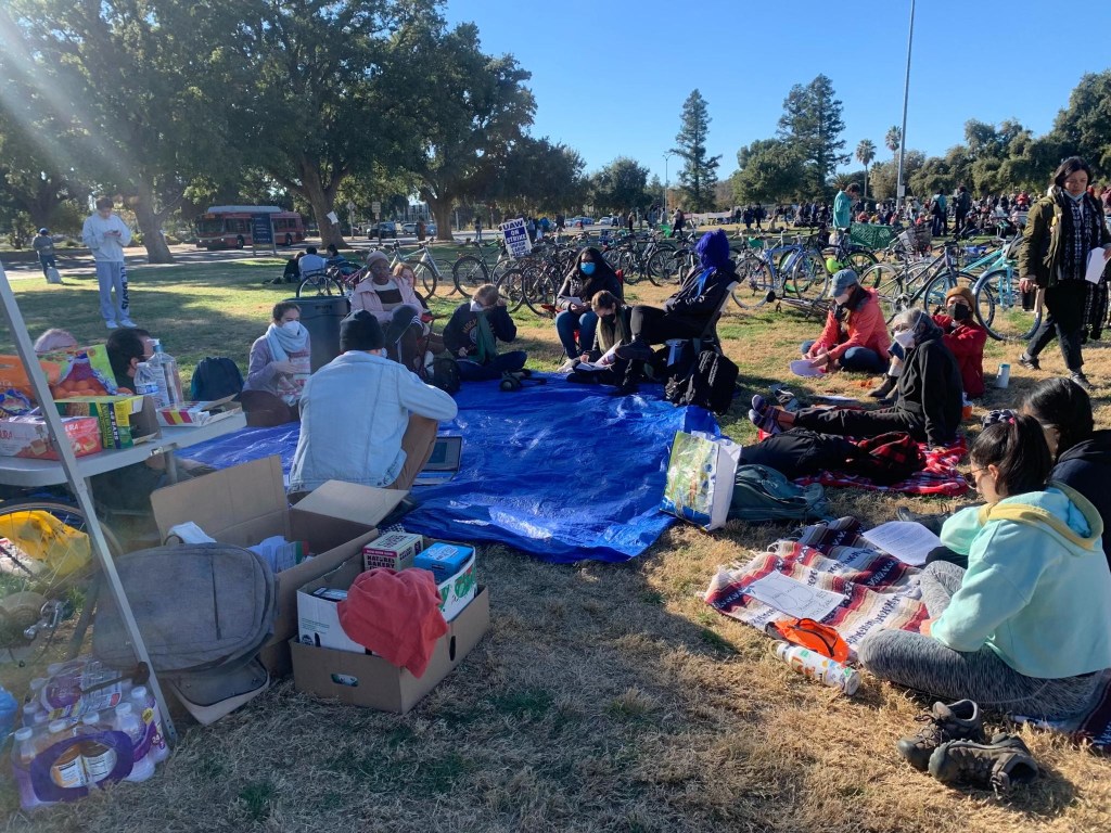 Stephen Eyman, with his back to the camera, sits on a blanket in front of the UC Davis mutual aid tent as he facilitates a disability studies teach-in. Next to him is a speaker so that those who cannot attend in-person can participate online. While most of the picketers in distance are not wearing masks, reflecting the general trend of strike, those circled around the blanket for the teach-in are almost all wearing masks and/or socially distanced. Photo of UC Davis Disability Studies Teach-In by Ingrid Sub Cuc. 2022. Used with permission.