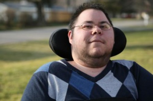 Photo of a man with short dark hair and glasses. He has stubble around his chin and neck. He is in a wheelchair with a black headrest behind him. He is wearing an argyle-print sweater in different shades of blue.