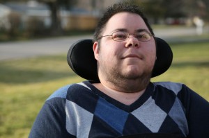 Photo of a man with short dark hair and glasses. He has stubble around his chin and neck. He is in a wheelchair with a black headrest behind him. He is wearing an argyle-print sweater in different shades of blue.