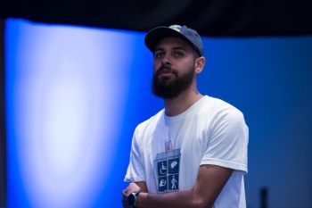 Image Description: Dustin, a Black man, stands in front of a blue backdrop with his arms folded looking out into an audience. He is wearing a dark blue hat and a white t-shirt with the #CripTheVote logo which is a 4 square box depicting different forms of disability.