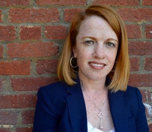 A woman with shoulder length red hair and green eyes standing against a brick wall. She is wearing a blue blazer and white t-shirt.