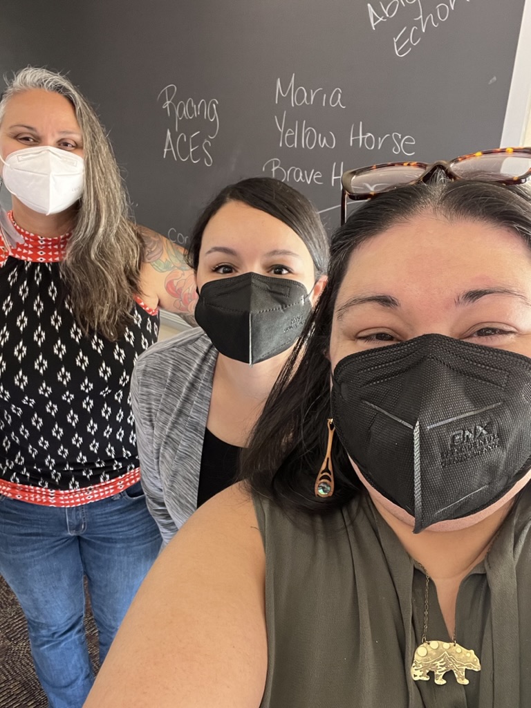 Una foto de tres miembros del personal de UPRISE COLLECTIVE. Brianna, a la derecha, es una nativa/Ihanktonwan que lleva máscara negra, camisa verde, collar dorado en forma de oso y pendientes de madera con conchas de abulón. En el centro está Stephanie, una mujer negra/nativa que lleva máscara negra y rebeca gris. Lleva el pelo negro recogido en la espalda. A la izquierda está Ginia, una persona Blauan, con máscara blanca, camisa multicolor y vaqueros. Todos posan delante de una pizarra.