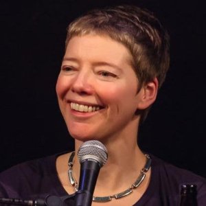 Photo against a black background of a white woman with short sandy blonde hair. She is smiling and looking slightly toward the left-hand side of the image. She has a metallic necklace on and a dark-colored top. A microphone is in front of her.