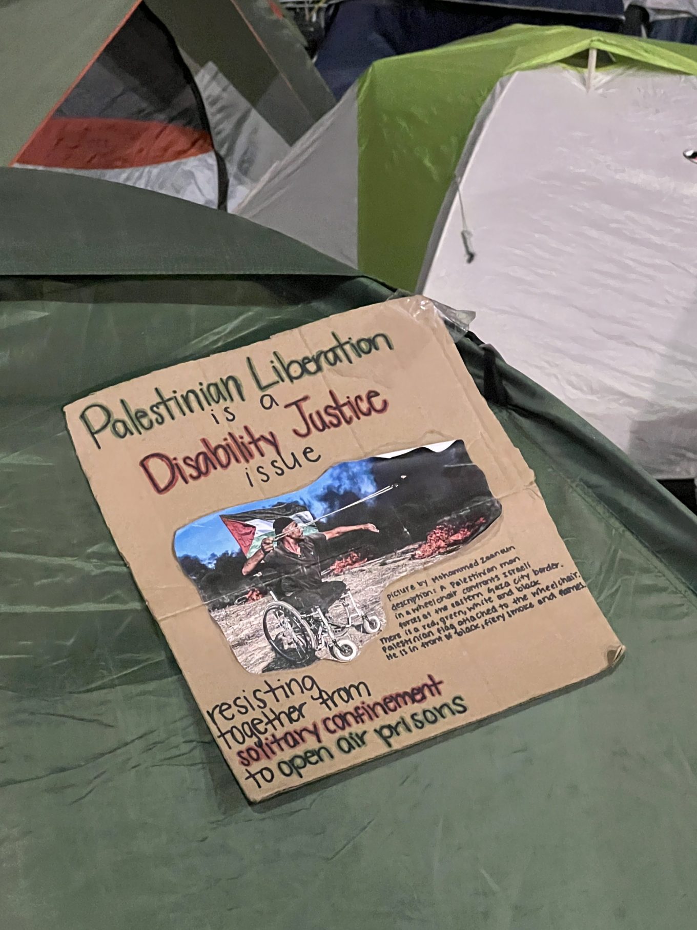 A cardboard sign on a tent at a student encampment in DC. It says at the top in red and black letters "Palestinian Liberation is a Disability Justice issue." In the center is a photo with the image description: "Picture by Mohammed Zaanoun" Description: A Palestinian man in a wheelchair confronts Israeli forces at the eastern Gaza City border. There is a red, green, white, and black Palestinian flag attached to the wheelchair. He is in front of black, fiery smoke and flames." Below the photo in black, red, and green lettering are the words "Resisting together from solitary confinement to open air prisons."