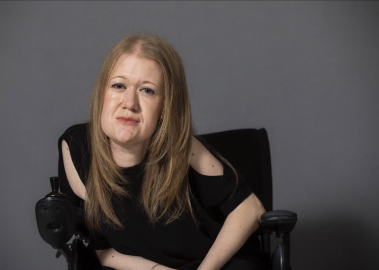 t 1: A photo of Frances Ryan, a white woman with blonde long hair sitting in a wheelchair. She is wearing a black top with subtle cut out details and is smiling.