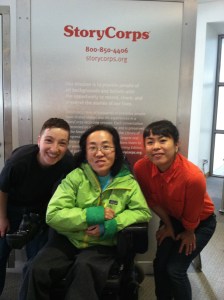 Asian American woman in a wheelchair in the middle of the image on her left and right are two other individuals, staff members of StoryCorps San Francisco. All three are in front of a metal StoryCorps recording booth