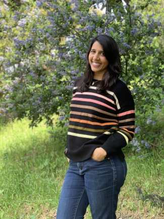 An Indian woman wearing a black sweater with pink and orange stripes and blue jeans smiles in front of a green tree with purple flowers. Her hands are in her pockets.