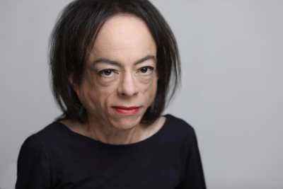 The photo shows the head and shoulders of a white woman, with make up and red lipstick, staring directly at you with her dark brown eyes. A slim woman, her navy blue long sleeved top frames her bare neck and her straight, dark brown hair frames her face and her high forehead.