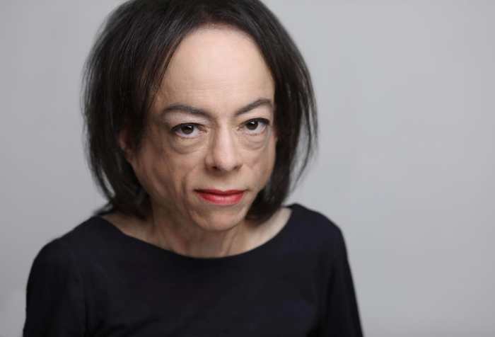 The photo shows the head and shoulders of a white woman, with make up and red lipstick, staring directly at you with her dark brown eyes. A slim woman, her navy blue long sleeved top frames her bare neck and her straight, dark brown hair frames her face and her high forehead.