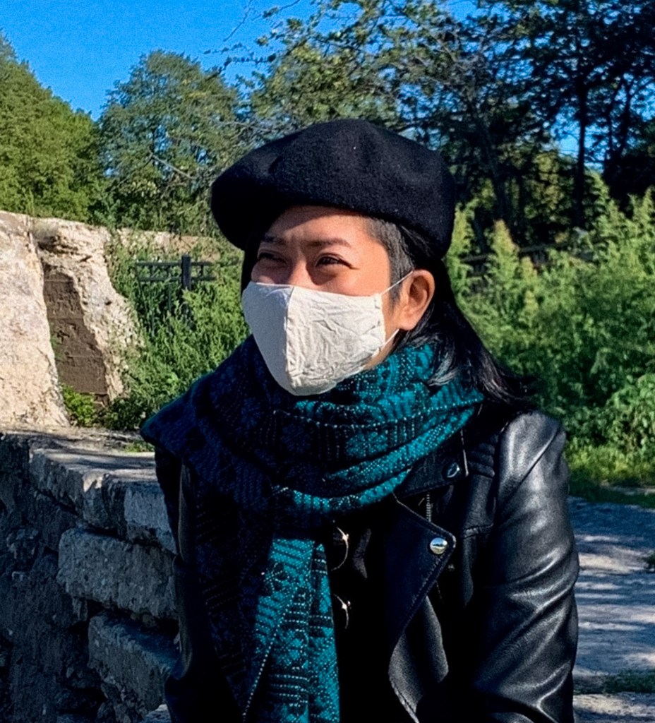 Foto de Akemi, mujer de Asia oriental con el pelo negro liso de longitud media rapado de lado. Lleva una boina negra, un pañuelo verde y una máscara blanca. Está sentada al aire libre frente al río y los árboles