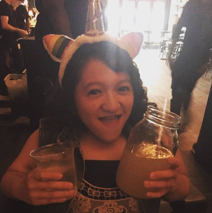 A photo of the author wearing a unicorn headband with rainbow ears and gold foil horn. She had dark shoulder length curly hair, is smiling with her mouth open. She has a cup of water in one hand and a mason jar with a straw in the other.