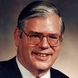 Older white man wearing a dark business jacket and a white shirt. He has glasses and is smiling at the camera.
