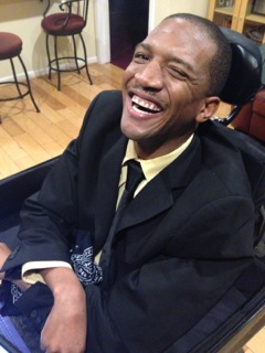African American man wearing a dark suit and a tie. He's sitting in a wheelchair and smiling at the camera.