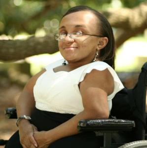 Photo of a young Black woman with her hair parted and hanging down straight.  She is smiling and facing the camera sideways in her wheelchair.  She is outside under the tree branches while wearing a white and black dress.  