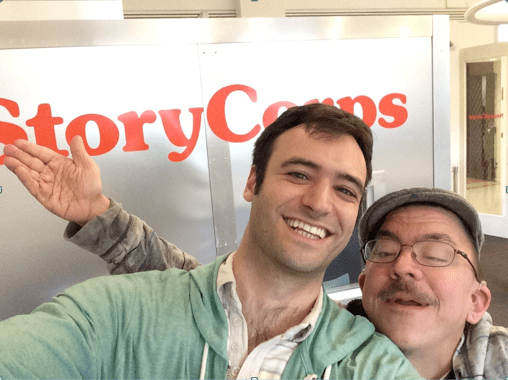Two men standing in front of a StoryCorps listening booth. The man on the left is younger with brown hair and brown eyes. The man on the right is older with glasses and a mustache. The arms of the second man are outstretched.